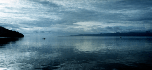 A fisherman in an outrigger canoe can be seen in a panoramic photo of the ocean with islands at dawn with an overcast sky.